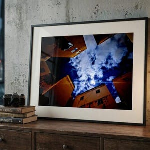 Puede incluir: Una impresión de arte mural horizontal enmarcada que muestra una perspectiva en contrapicado de edificios naranjas que rodean un cielo nocturno azul oscuro con nubes brillantes y una luna luminosa. Marco negro con paspartú blanco.