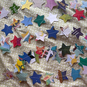 May include: A decorative garland featuring numerous five-pointed stars cut from colorful printed paper and book pages. The stars are strung together on a thin white thread, displaying various illustrations, text, and solid colors in a multi-colored arrangement.
