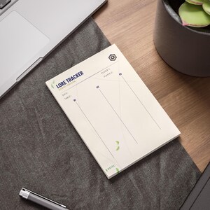 Puede incluir: Un bloc de notas de papel rectangular titulado LORE TRACKER DISNEY LORCANA en texto azul oscuro. La hoja de color blanco hueso presenta campos para Player 1, Player 2, Date y Table, con columnas verticales etiquetadas R1, R2 y R3, y notas.