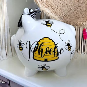 May include: White ceramic piggy bank with a black and white striped bow. The bank features a yellow beehive design with the name "Phoebe" in black script, and several cartoon bees with dotted flight paths.