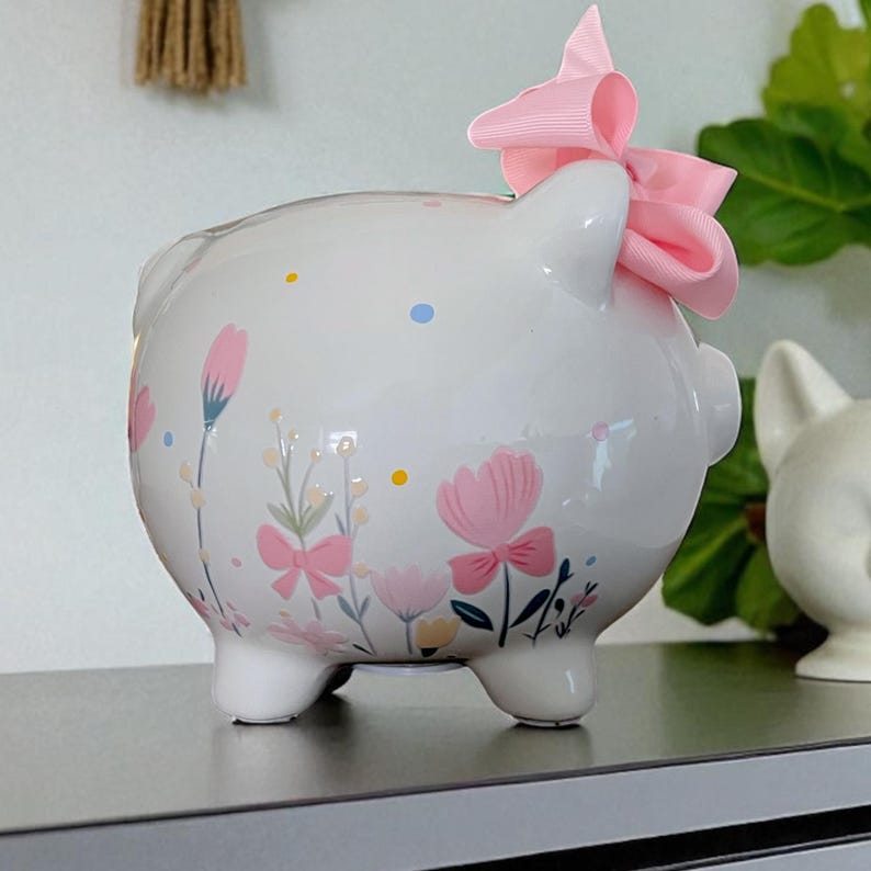 以下が含まれることがあります： ピンクの花、緑の茎、小さな黄色と青のドットが描かれた白い陶器製の貯金箱。貯金箱の上部、耳の近くには、淡いピンク色のグログランリボンの大きな結び目が付いています。