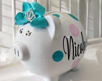 May include: White ceramic piggy bank with turquoise and pink polka dots. The piggy bank has a turquoise bow with a silver crown on top and the name "Nicole" written in black.