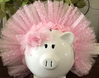 May include: White ceramic piggy bank with black eyes and nostrils, wearing a pink tulle tutu with white polka dots and a pink flower crown.