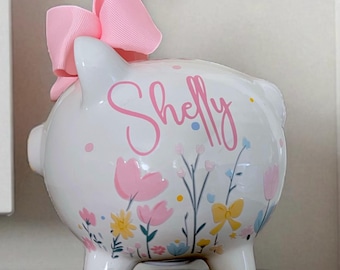 May include: A white ceramic piggy bank featuring a large pink ribbon bow on top. The side is decorated with colorful painted flowers in shades of pink, yellow, and blue, along with the name Shelly written in a cursive pink script font.