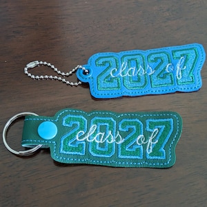 May include: Two rectangular keychains featuring the embroidered text "class of 2027". The top keychain is light blue with dark green numbers and a silver ball chain. The bottom keychain is dark green with light blue numbers, a snap, and a ring.