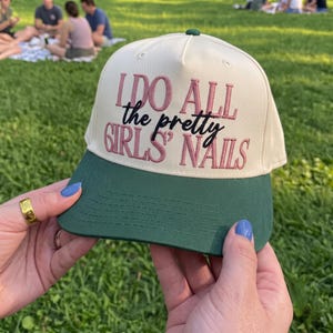 May include: A cream-colored baseball cap with a dark green brim. The front panel features embroidered text in pink and black that reads: I DO ALL the pretty GIRLS' NAILS. The cap has a structured crown and a button on the top center.