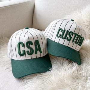 May include: Two baseball caps featuring a white base with thin vertical black pinstripes and a solid dark green brim. One cap displays the green raised letters CSA on the front, while the other cap features the green raised text CUSTOM.