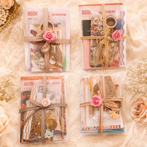 May include: Four rectangular clear plastic packages containing assorted stationery items, including paper sheets, stickers, and decorative cutouts. Each package is tied with a ribbon and adorned with a small pink fabric rose flower accent.