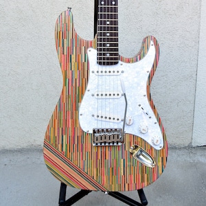May include: A colorful electric guitar with a white pickguard and a black fretboard. The body of the guitar is made of recycled skateboard decks, creating a unique and eye-catching design. The guitar has a "S-Strings" logo on the headstock.