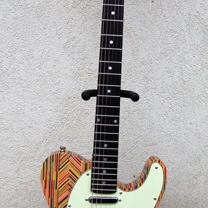 May include: A unique electric guitar with a body made from colorful, vertically aligned wood strips. The guitar features a light green pickguard, black hardware, and a natural wood neck with the brand name "Skarings" on the headstock.