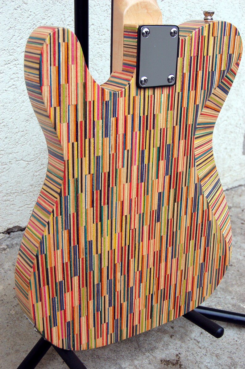 May include: A colorful electric guitar with a body made from recycled skateboard decks. The back of the guitar features a vertical striped pattern of various colors, including red, yellow, blue, and green. The neck is made of light-colored wood.