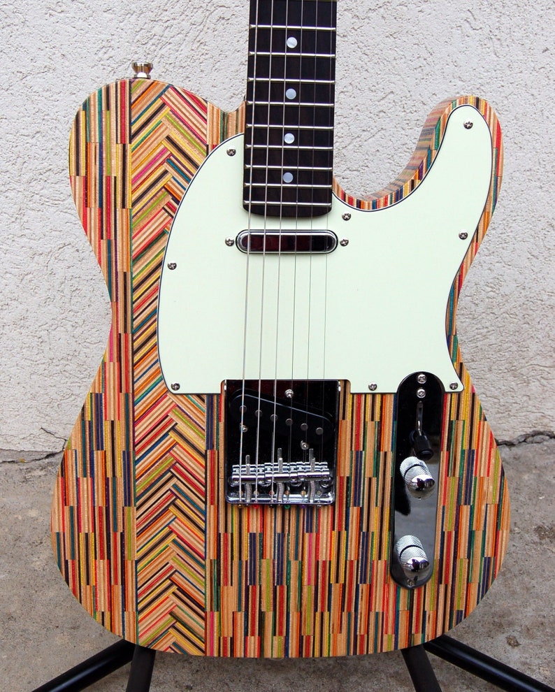 May include: A colorful electric guitar with a unique body made from vertical strips of wood in various colors. The guitar has a white pickguard, black pickups, and chrome hardware. The fretboard is black with white inlays. The guitar is on a black stand.