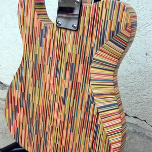 May include: A colorful electric guitar with a body made from layered wood strips in various colors. The back of the guitar is visible, showing the neck joint, metal plate, and tuning peg. The guitar is on a black stand.