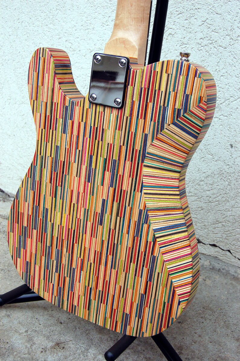 May include: A colorful electric guitar with a body made from layered wood strips in various colors. The back of the guitar is visible, showing the neck joint, metal plate, and tuning peg. The guitar is on a black stand.