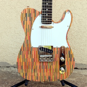 Puede incluir: Una guitarra eléctrica colorida con una golpeador blanco y un cuerpo de madera hecho de tablas de skate recicladas. La guitarra tiene una pastilla de bobina simple y un selector de tres vías.
