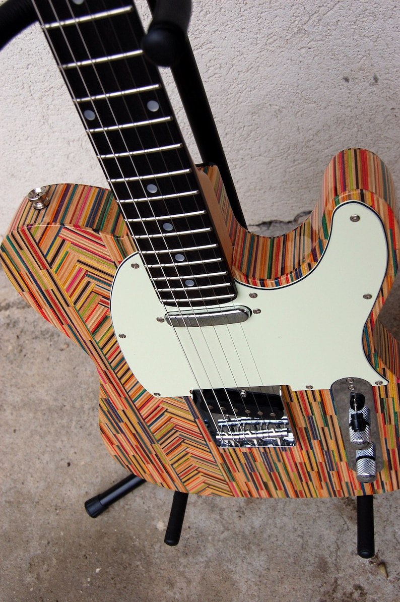May include: A unique electric guitar with a body crafted from colorful, layered wood, possibly repurposed skateboard decks. The guitar features a white pickguard, chrome hardware, and a black neck with frets. It rests on a black stand.