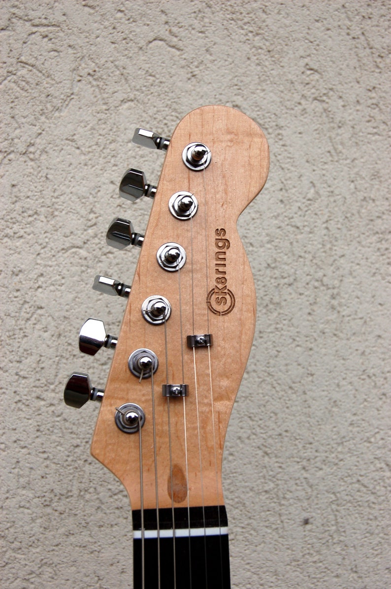 May include: Close-up of a guitar headstock with chrome tuning pegs and strings. The wood is light-colored with the brand name "Sk8rings" engraved on the headstock. The fretboard is black and white.