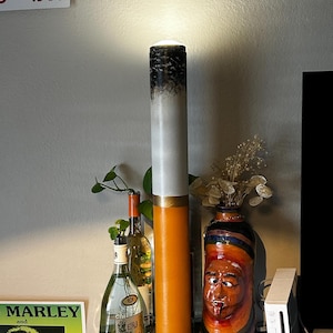May include: A tall, cylindrical lamp designed to resemble a lit cigarette, featuring a white body with a black top and an orange filter section at the base. The lamp emits a soft glow from the top, standing upright on a small, square, white base.