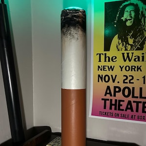 May include: A tall, cylindrical lamp shaped like a lit cigarette, featuring a brown filter section at the base, a white paper middle, and a textured, dark gray ash top that emits a soft green light from the center of the glowing tip.