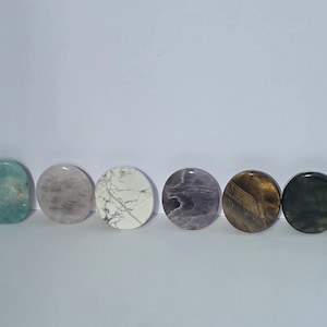 May include: Eight circular polished stones arranged in a horizontal row. The stones feature various colors and patterns, including solid reddish-brown, light blue, pale grey, white with grey veining, purple, brown tiger's eye, dark green, and mottled green and purple.