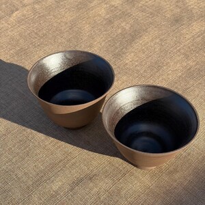 Puede incluir: Dos cuencos de cerámica con un exterior marrón mate y un interior negro liso. Los cuencos presentan un borde ancho y abierto que se estrecha hacia una base pequeña, mostrando un contraste marcado entre la superficie exterior terrosa y el esmalte oscuro.