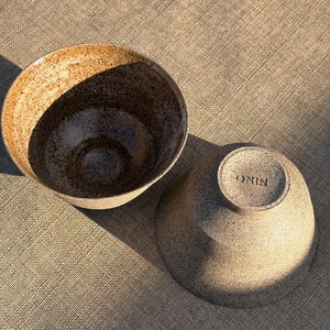 Puede incluir: Dos cuencos de cerámica con un acabado moteado en tonos bronceado y marrón. Un cuenco está colocado boca arriba, revelando un interior oscuro y texturizado, mientras que el otro está invertido, mostrando una base lisa con la palabra ONIN.