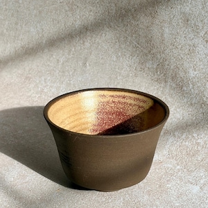 Puede incluir: Un pequeño cuenco de cerámica redondo con un exterior de color marrón oscuro mate y un interior en contraste que presenta un acabado dorado metálico con motas de color marrón rojizo. El cuenco tiene un borde ancho y una silueta cónica.