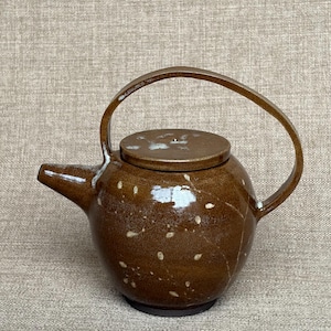 Puede incluir: Una tetera de cerámica marrón con acabado brillante y un asa alta y arqueada. El cuerpo presenta un patrón moteado de pequeños puntos blancos y una tapa circular a juego. El pico se extiende por el lateral y la base es de tono marrón mate.