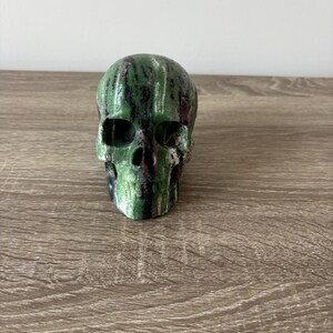 Puede incluir: Una escultura de cráneo tallada en piedra que presenta una mezcla vibrante de inclusiones minerales en verde oscuro, negro y rosa magenta. La superficie muestra un acabado pulido con vetas verticales naturales y cuencas oculares definidas.