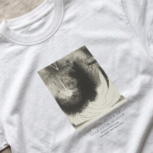 May include: A white short-sleeved t-shirt featuring a rectangular black and white graphic print on the chest. The graphic depicts a dark, swirling abstract form. Below the image, the text reads: AND THERE FELL A GREAT STAR FROM HEAVEN.