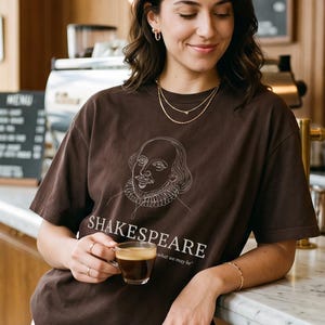 Op de afbeelding: Een donkerbruin t-shirt met korte mouwen voorzien van een witte lijntekening van het gezicht van William Shakespeare, de tekst "SHAKESPEARE" in een schreeflettertype en kleinere tekst eronder: "to be or not what we may be".