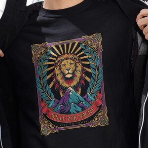 Könnte beinhalten: Ein schwarzes T-Shirt mit einem Grafikdruck eines Löwen über einem Gebirge, eingerahmt von einer verzierten Bordüre mit Lorbeerblättern. Das Design enthält ein rotes Banner mit dem Text "WILLE ZUR MACHT" und "WILL TO POWER" in Weiß.