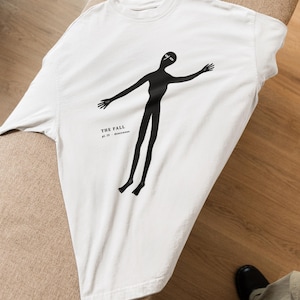 May include: A white crew neck t-shirt featuring a black graphic of a stylized, elongated human figure with arms outstretched. The text "THE FALL" and "pl. IV - descensus" is printed in black lettering to the left of the figure's torso.