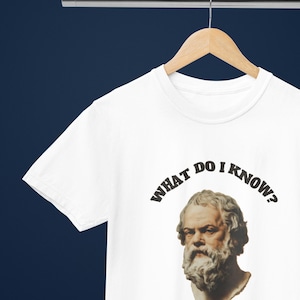Könnte beinhalten: Ein weißes kurzärmeliges T-Shirt mit einem Grafikdruck einer klassischen Büste von Sokrates. Über der Büste ist der Text "WHAT DO I KNOW?" in einer fetten, dunklen Serifenschrift gedruckt. Das T-Shirt hängt an einem Holzkleiderbügel vor dunklem Hintergrund.