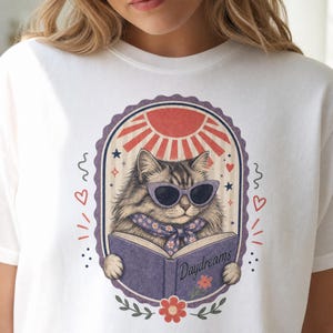 Puede incluir: Una camiseta blanca con el gráfico de un gato esponjoso que lleva gafas de sol oscuras y un pañuelo floral, leyendo un libro morado con la palabra Daydreams. El diseño incluye un sol rojo, estrellas pequeñas, corazones y flores.