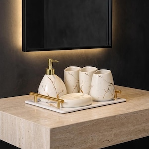 Op de afbeelding: Een badkameraccessoireset bestaande uit een witte zeepdispenser met een gouden pomp, twee bijpassende bekers en een zeepbakje, allemaal versierd met een goudkleurig marmerpatroon. De set staat op een rechthoekig wit dienblad met gouden handgrepen.