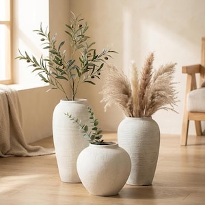 May include: Three off-white ceramic vases with a textured, matte finish. The tallest vase holds green olive branches, the medium vase contains fluffy beige pampas grass, and the smallest, round vase holds a single sprig of green eucalyptus leaves.
