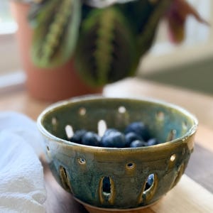 May include: A round, glazed ceramic berry bowl featuring a mottled green and blue finish with a series of vertical and circular drainage holes around the sides. The bowl is filled with a small handful of fresh, dark blue blueberries.