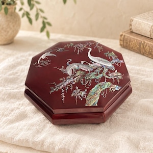 May include: A hexagonal dark red lacquered box featuring an intricate mother-of-pearl inlay design on the lid. The inlay depicts three cranes standing among pine branches and foliage, with a fourth crane in flight against a dark background.