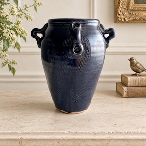 May include: A deep navy blue ceramic vessel with a glossy finish and a textured, diagonal ribbed pattern. The jar features two prominent, curved handles on opposite sides and a wide, open mouth, with a visible light-colored clay base.