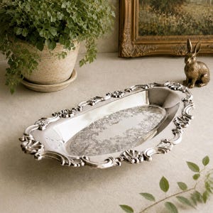 May include: An oval silver-toned serving tray featuring a polished, reflective surface with an intricate, embossed floral pattern in the centre and a decorative, scalloped border with shell-like motifs along the entire outer rim.