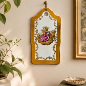 May include: A rectangular ceramic wall plaque with a white centre featuring a colourful illustration of two figures in a landscape. The plaque has a gold-coloured border with a delicate leaf pattern and a circular hole at the top for hanging.