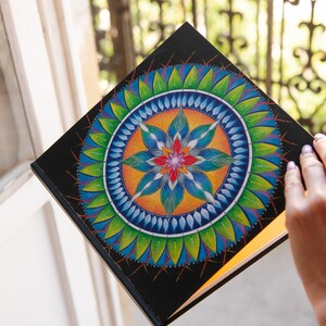 May include: A black journal with a colorful mandala design on the cover. The mandala features a central flower with blue, green, orange, and red petals surrounded by concentric circles of leaves and dots.