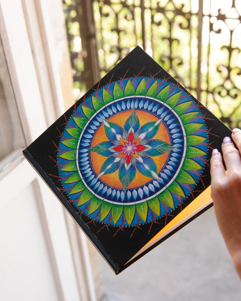 May include: A black journal with a colorful mandala design on the cover. The mandala features a central flower with blue, green, orange, and red petals surrounded by concentric circles of leaves and dots.