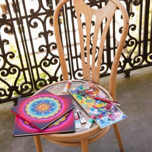 May include: Two colorful mandala art journals with red, yellow, blue, and green colors, a paint palette with various colors of paint, and paint tubes on a wooden chair.