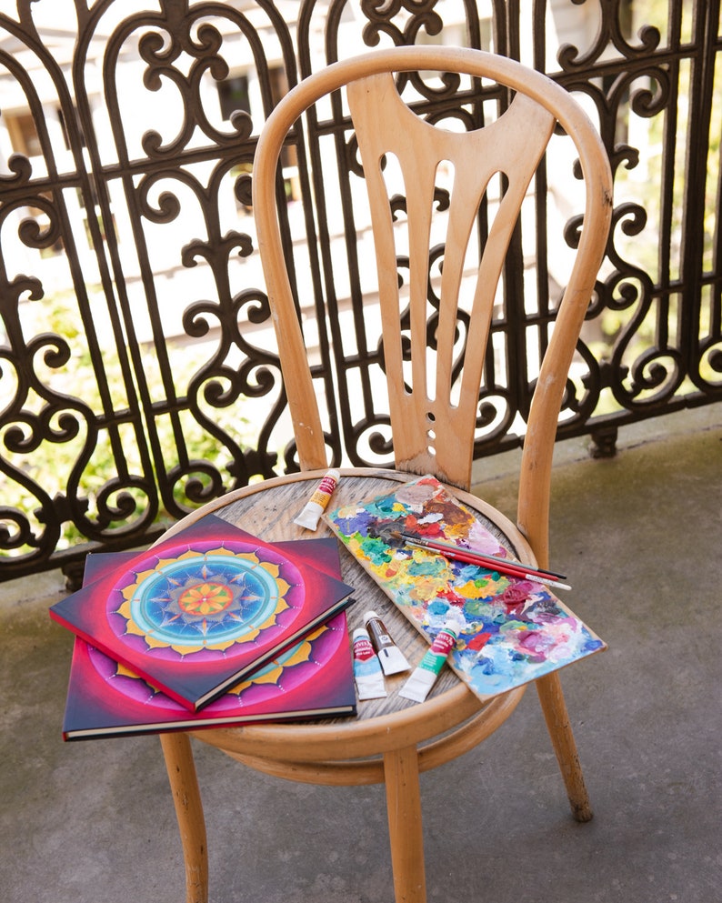 May include: Two colorful mandala art journals with red, yellow, blue, and green colors, a paint palette with various colors of paint, and paint tubes on a wooden chair.
