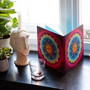 May include: A closed book with a colorful mandala design on the cover. The book is sitting on a windowsill with a lit candle and potted plants.
