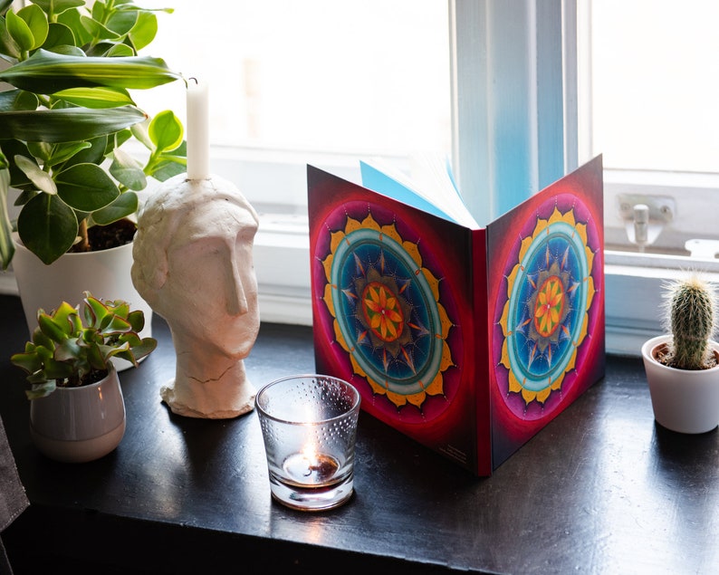 May include: A closed book with a colorful mandala design on the cover. The book is sitting on a windowsill with a lit candle and potted plants.