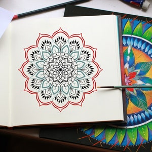 May include: A black and white mandala design with red accents is drawn in a sketchbook. A paintbrush is lying on the page. A second mandala design is painted in vibrant colors on a black canvas.