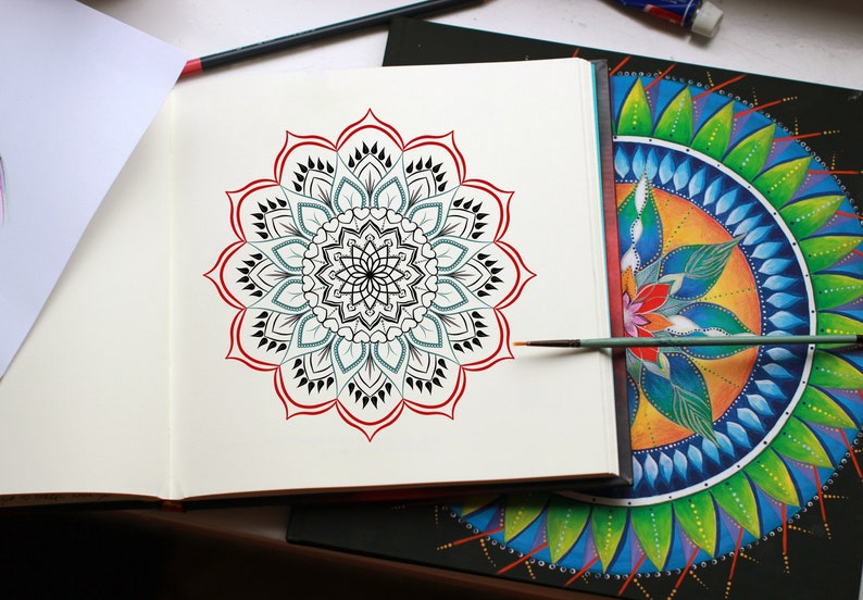 May include: A black and white mandala design with red accents is drawn in a sketchbook. A paintbrush is lying on the page. A second mandala design is painted in vibrant colors on a black canvas.